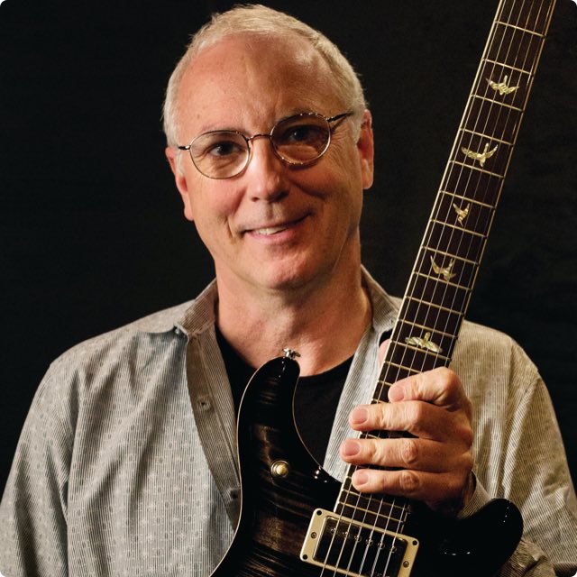 Paul Reed Smith in front of a black background holding out a guitar
